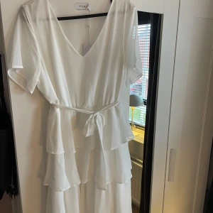 Vit omlottklänning med volang från Vila - Superfin vit klänning från Vila med v-ringning, volangdetaljer och knytband i midjan. Klänningen har korta ärmar och är tillverkad i ett lätt, flowy material. Klänningen är aldrig använd och har prislappen kvar. 
