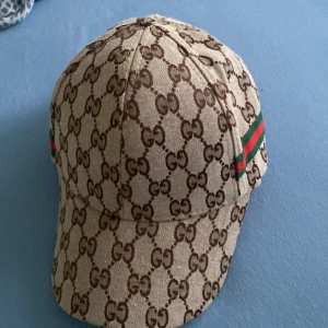 Beige keps från Gucci med GG-mönster - Säljer en beige keps från Gucci med klassiskt brunt GG-mönster över hela kepsen och en grön-röd rand på sidan. Perfekt accessoar för att lyfta din outfit. Priset kan diskuteras.aldrig använd 