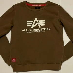 Brun sweatshirt Alpha Industries - Säljer en brun sweatshirt från Alpha Industries med vit logga och text på bröstet. Tröjan har rund halsringning, långa ärmar och ribbade muddar vid ärmslut och nederkant. Perfekt för en avslappnad stil.