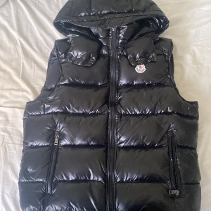 Svart dunväst från Moncler med huva - Säljer en svart, glansig dunväst från Moncler med avtagbar huva och dragkedja framtill. Västen har två fickor med dragkedja och klassisk Moncler-logga på bröstet. Har använts bara 3 gånger, den är helt sprillans ny kan man säga