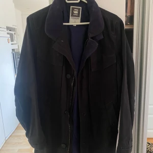 Svart kappa  från G-Star Raw - Säljer en svart jacka från G-Star Raw med klassisk krage och knäppning framtill. Jackan har två snedställda fickor och en mjuk insida vid kragen. Perfekt för dig som vill ha en stilren och tidlös look.