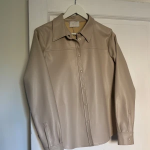 Beige jacka i skinnimitation från Neo Noir - Snygg beige jacka från Neo Noir i skinnimitation. Skjortan har klassisk krage, knäppning framtill och långa ärmar med manschett. Perfekt för dig som gillar stilrena och trendiga plagg i neutrala färger.