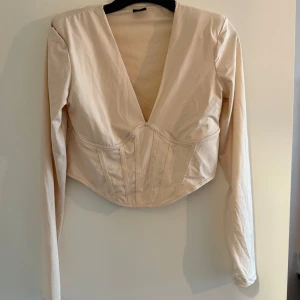 Beige långärmad korsettopp - Snygg beige korsettopp med lång ärm och djup v-ringning. Toppen har markerad midja och croppad passform som ger en trendig siluett. Perfekt att styla till jeans eller kjol