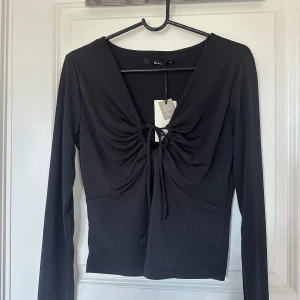 Svart långärmad topp med knyt från Bik Bok - Säljer en svart långärmad topp från Bik Bok med v-ringning och snygg knytdetalj framtill. Toppen har en figurnära passform och är perfekt att styla till olika outfits.