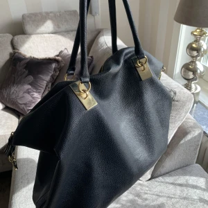 Svart handväska med gulddetaljer - Stilren svart handväska i skinnimitation med två handtag och guldfärgade metalldetaljer. Väskan har dragkedja upptill och ett innerfack med dragkedja. Perfekt för dig som vill ha en rymlig och klassisk accessoar.