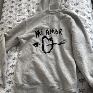 Mi Amor grå hoodie från Mira Paris - Grå hoodie från Mira Paris med svart tryck på ryggen där det står 'Mi Amor' och ett hjärta genomborrat av en pil. Klassisk känguruficka framtill och justerbar huva med dragsko. Perfekt för en avslappnad stil.