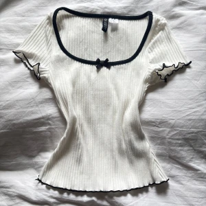 Vit ribbad topp med svarta detaljer och rosett - Supersöt vit ribbad topp med svarta kanter och liten rosett framtill. Toppen har korta ärmar med vågig kant och en fyrkantig urringning. Perfekt för en trendig och feminin look.