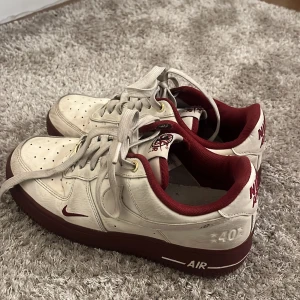 Nike Air Force 1 i vitt och vinrött - Säljer ett par Nike Air Force 1 med vit ovandel och vinröd sula samt detaljer. Skorna har klassisk Nike-logga på sidan, röd insida och texten NIKE AIR broderad bak. Perfekta för dig som gillar retrostil och streetwear.