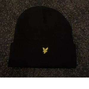 Svart mössa från Lyle & Scott - Svart mössa från Lyle & Scott med gul broderad logga framtill. Klassisk modell med uppvikt kant, perfekt för kyliga dagar. Enkel och stilren design som passar till det mesta.
