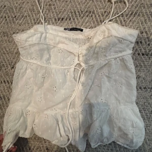 Vit broderad linnetopp med knyt - Söt vit linnetopp från Zara med tunna axelband och broderade blommor. Toppen har knyt framtill och volang nertill för en romantisk känsla. Perfekt för varma dagar!