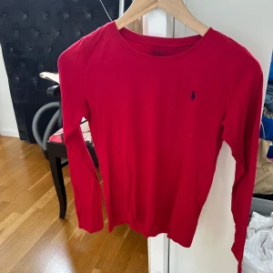 Röd långärmad tröja från Polo Ralph Lauren - Säljer en klassisk röd långärmad tröja från Polo Ralph Lauren med rund halsringning och den ikoniska blå loggan broderad på bröstet. Perfekt till jeans eller kjol för en stilren look. (Barn storlek xl, 12/14 men passar som dam xs)