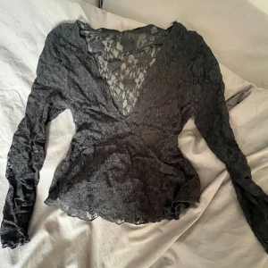 Svart spetsblus med v-ringning - Säljer en svart blus i genombruten spets med blommönster och djup v-ringning. Långärmad och figurnära passform, perfekt för att skapa en snygg och trendig look.