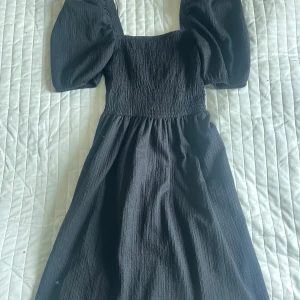 Svart klänning med puffärmar - Säljer en svart klänning med markerad midja och stora puffärmar. Klänningen har en fyrkantig ringning och är tillverkad i ett strukturerat tyg som ger en snygg siluett. Perfekt för dig som gillar trendiga detaljer och klassisk stil.