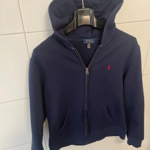 Mörkblå hoodie från Ralph Lauren - Mörkblå hoodie från  Ralph Lauren med dragkedja framtill och klassisk röd logga på bröstet. Tröjan har huva och två fickor framtill, perfekt för en avslappnad stil. Storlek xl i barn vilket motsvarar S. Vid frågor är det bara att höra av dig.