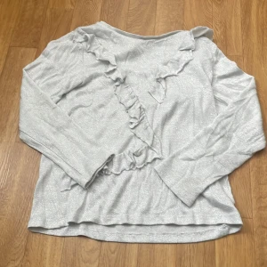Ljusgrå glittrig tröja med volang från Dreams - Säljer en ljusgrå glittrig tröja från Dreams i storlek 140. Tröjan har långa ärmar och dekorativa volanger framtill som ger en snygg detalj. Perfekt för dig som gillar lite extra glitter i vardagen!