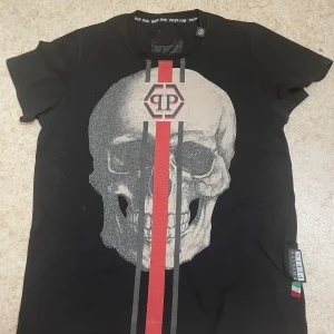 Svart t-shirt med dödskalletryck från Philipp Plein - Cool svart t-shirt från Philipp Plein med stort dödskalletryck framtill och röda samt gråa ränder. Rund halsringning och korta ärmar. Perfekt för dig som gillar edgy streetstyle.