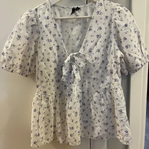 Vit blommig blus med knyt fram - Supersöt vit blus med blått blommönster, puffiga korta ärmar och v-ringning med knyt fram. Blusen har en luftig passform och volang nedtill, perfekt för en fräsch och romantisk look. Tillverkad i mjuk bomull som känns skön mot huden.