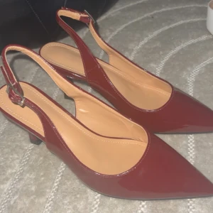 Helt nya Vinröda pumps, (kommer med matchande väska) - HELT OANVÄNDA, Snygga vinröda pumps med lackad finish och spetsig tå. Skorna har öppen häl med justerbart spänne och en smal klack som ger en elegant look. Perfekta för dig som vill sticka ut med stilrena detaljer och trendig färg, priset går att diskuteras vid snabb affär! Kommer med matchande väska!