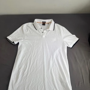 Vit pikétröja från BOSS med svarta detaljer - Vit pikétröja från BOSS med klassisk krage och svarta ränder på ärmslut och krage. Tröjan har korta ärmar, knäppning framtill och diskret BOSS-logga på bröstet. Tillverkad i mjuk bomull som känns skön mot huden.