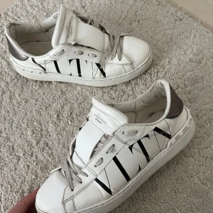 Valentino open - Snygga vita sneakers från Valentino Garavani med svart VLTN-logga på sidan och silverfärgad detalj vid hälen. Skorna har snörning, platt sula och är tillverkade i skinn. Perfekta för dig som gillar exklusiva och trendiga sneakers. Strl 41,5 priset är diskuterbart