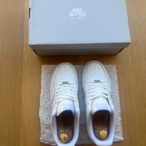 Nike Air Force 1 helvita sneakers - Klassiska Nike Air Force 1 sneakers i helvitt skinn med vit sula och snörning. Snygg och tidlös modell med perforerad tå och ikonisk swoosh på sidan. Perfekta för dig som vill ha en clean och fräsch look till vardags. Levereras med originalkartong.