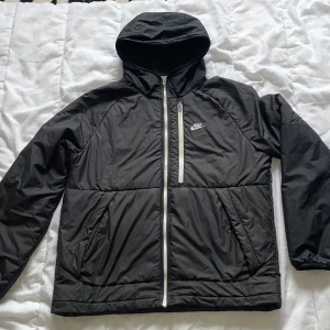 Svart Nike vindjacka med huva L - Svart vindjacka från Nike med Therma-Fit-teknologi. Jackan har huva, hel dragkedja framtill och två sidofickor. Snygg vit Nike-logga på bröstet och kontrasterande vit dragkedja. Perfekt för kyliga dagar och sportiga outfits.