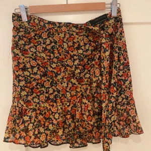 Blommig omlottkjol Urban Outfitters - Supersöt kort omlottkjol från Urban Outfitters med blommigt mönster i orange, gul och röd på svart botten. Kjolen har volangkant och knyt i sidan! 