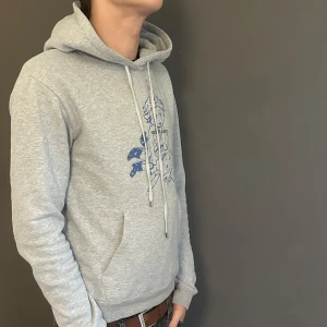 Grå Saint Laurent hoodie med print - Grå hoodie från Saint Laurent med blå tecknat print framtill och klassisk känguruficka. Tröjan har huva med vita snören och ribbade muddar. Perfekt för dig som gillar streetstyle och vill ha något unikt från ett lyxmärke.