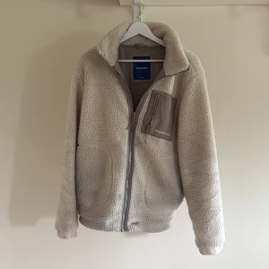 Beige teddyjacka från Jack & Jones - Mysig beige teddyjacka från Jack & Jones Originals med hel dragkedja och hög krage. Perfekt för kalla sommar kvällar där du vill vara varm och stilren på samma gång! Hör av dig vid minsta funderingar?