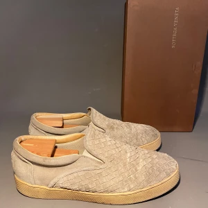 Beige slip-on sneakers från Bottega Veneta - Snygga slip-on sneakers från Bottega Veneta i beige mocka med deras ikoniska flätade mönster. Skorna har en platt sula i gummi och rund tå. Perfekta för dig som gillar stilrena och exklusiva detaljer.