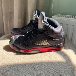 Nike Air Jordan 5 Retro svart/röd - Säljer ett par Nike Air Jordan 5 Retro med svart ovandel, grå plös med röd Jordan-logga och detaljer i rött på sulan. Skorna har snörning, meshpaneler på sidorna och genomskinlig yttersula. Perfekta för dig som gillar klassiska sneakers med streetkänsla men ändå vill sticka ut .