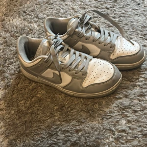 Nike Dunk Low i grått och vitt - Nike Dunk Low sneakers i stilren grå och vit färgkombination. Klassisk siluett med rund tå, snörning och platt sula. Ovandel i skinn med perforerade detaljer på tån för extra ventilation. Perfekta för dig som gillar en clean och tidlös look.