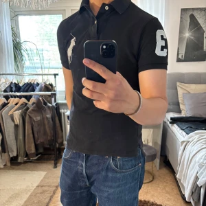 Svart pikétröja Polo Ralph Lauren S - Svart pikétröja från Polo Ralph Lauren med klassisk krage, två knappar och stor vit broderad logga på bröstet. Snygg detalj med siffran 3 på ärmen. Tillverkad i bomull och har korta ärmar. Perfekt för en clean och sportig look. (Notering) Har egentligen storlek M så den sitter lite tajt på mig