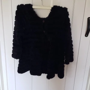 Svart fluffig pälsjacka - Mysig svart pälsjacka med fluffig känsla och rund hals. Jackan har knappar framtill och trekvartsärmar. Perfekt för dig som vill ha en statementjacka med skön vibe och extra volym. Passar till lager-på-lager och ger en cool look.