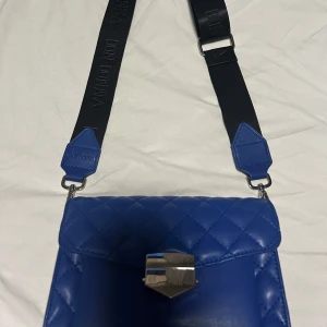 Blå axelväska från Don Donna - Blå axelväska från Don Donna och bred svart axelrem med logga. Väskan har silvrigt spänne framtill.