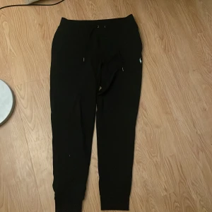 Svarta mjukisbyxor Polo Ralph Lauren - Svarta mjukisbyxor från Polo Ralph Lauren med dragsko i midjan och broderad logga på ena benet. Byxorna har ribbade muddar vid bensluten och är gjorda i en mjuk bomulls- och polyestermix. Perfekta för chill eller träning.