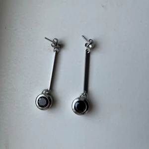 Silvriga örhängen från Edblad - Snygga silvriga örhängen från Edblad med raka stavar och rund svart sten i botten. Modern och stilren design, perfekta för att lyfta din look. Märket är ingraverat på baksidan av örhängena. Passar dig som gillar minimalistiska smycken. Aldrig använd.