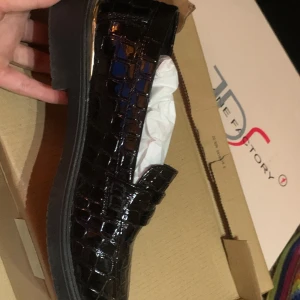 Svarta loafers med krokopräglat mönster - Snygga svarta loafers med glansig krokopräglad yta och platt sula. Skorna har en klassisk form och en liten gulddetalj vid hälen som ger en extra touch. Perfekta för dig som vill ha en stilren men ändå trendig look.