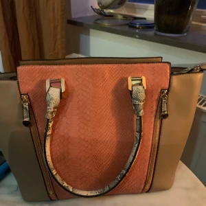 Beige och rosa handväska med ormmönster - Snygg handväska i beige och rosa med ormmönstrade handtag och detaljer. Väskan har strukturmönstrad framsida, guldfärgade metalldetaljer och dragkedjor på sidorna. Rymligt innerfack med flera småfack för mobilen och andra prylar. Perfekt för dig som gillar unika accessoarer.