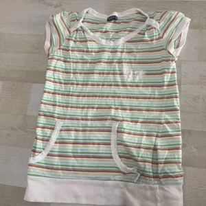 Randig topp med fickor och knappar - Söt randig topp från Kappahl i vitt med gröna, gula, rosa och blå ränder. Toppen har korta ärmar, två fickor framtill och dekorativa knappar vid axeln. 