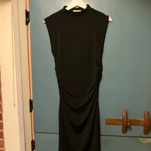 Svart långklänning med drapering - Stilren svart långklänning med hög hals och ärmlös design. Klänningen har snygg drapering längs sidorna som ger en cool siluett. Tillverkad i ett mjukt, stretchigt material som sitter bekvämt och följsamt. Perfekt för dig som gillar minimalistisk och modern stil.