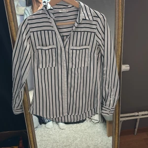 Vit och svart randig skjorta - Vit skjorta med svarta vertikala ränder, klassisk krage och två bröstfickor. Långärmad med knappar vid ärmslut och ärmslejfar. Skjortan har en rak passform och är tillverkad i ett lätt och mjukt material som känns skönt mot huden.