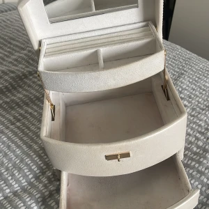Vit smyckesväska med spegel - Snygg oval smyckesväska i vit skinnimitation med guldfärgade detaljer och praktiskt handtag. Väskan har flera fack, spegel på insidan och smart lås framtill. Perfekt för att organisera smycken och accessoarer.