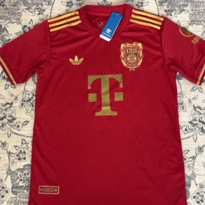 FC Bayern München 125 år Adidas tröja - Röd FC Bayern München 125-årsjubileumströja från Adidas i slim fit. Guldiga detaljer som klubbmärke, Adidas-logga, sponsortryck och axelränder. Tillverkad i lätt och ventilerande material, perfekt för match eller träning. Klassisk rund hals och korta ärmar.
