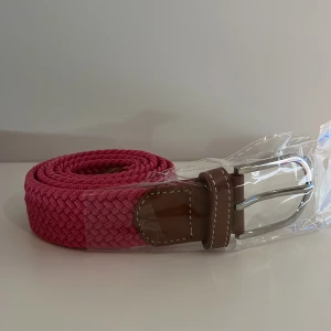 Flätat rosa bälte  - Snyggt flätat bälte i stark rosa färg med bruna skinnliknande detaljer och silvrigt spänne. Perfekt accessoar för att lyfta din outfit med lite färg. Bältet har en klassisk design med vit kontrastsöm på läderdetaljerna.