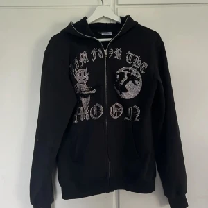 Svart hoodie med strass och tryck - En svart hoodie med dragkedja från Aim for the Moon som inte säljs längre. Alla stenar är kvar och den har nästan inte används någonting, orginalpris är 750kr💕första bilden är lånad