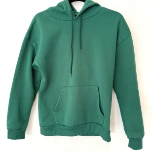 Grön hoodie med huva och ficka - Enkel grön hoodie i storlek S med huva och dragsnören. Tröjan har en stor magficka framtill och ribbade muddar vid ärmslut och nederkant. Perfekt för chill dagar eller när du vill ha en avslappnad stil.