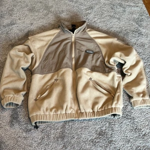 Beige fleecejacka från iets frans... - Säljer en beige fleecejacka från iets frans... med kontrasterande ljusbruna paneler och broderad logga på bröstet. Jackan har dragkedja framtill, två snedställda fickor med dragkedja och elastiska muddar. Perfekt för lager-på-lager och chill vardagsstil.