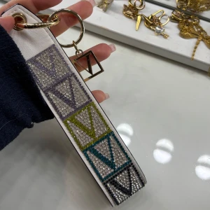 Vit nyckelring/strap från Victoria’s Secret - Säljer en vit strap/nyckelring från Victoria’s Secret med glittrande stenar i lila, gul, blå och svart. Remmen har ett stort V-mönster i olika färger och en guldfärgad ring med Victoria’s Secret-logga. Perfekt som accessoar till väska eller nycklar.