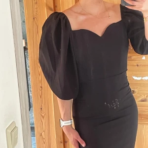 Svart kort klänning med puffärm - Supersnygg svart kort klänning med puffiga trekvartsärmar och hjärtformad urringning. Tight passform som framhäver figuren och öppen rygg för en extra cool look. Perfekt för dig som vill sticka ut med en elegant men edgy stil.